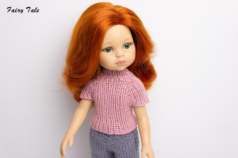 Pode incluir: Uma boneca com cabelo comprido e encaracolado vermelho usando um su&eacute;ter de tric&ocirc; rosa e cal&ccedil;as cinza. A boneca tem olhos verdes e uma tez clara.