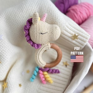 以下が含まれることがあります： 木製のリングが付いた、かぎ針編みのユニコーンのベビーラトル。ユニコーンはクリーム色の体、紫色のたてがみ、ピンクの角をしています。ラトルは虹色の尾を持っています。金色の星のアクセントが付いた白い背景。
