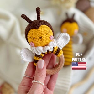 May include: A crocheted bee rattle toy with a wooden ring. The bee is yellow and brown with white wings and a white ruffle collar. The bee has a cute face with closed eyes and pink cheeks. The rattle is a great toy for babies and toddlers.