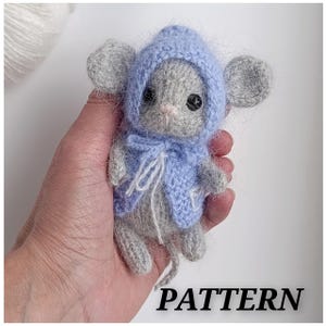 May include: A handmade gray and blue crocheted mouse wearing a blue hooded jacket. The mouse has large gray ears, black eyes, and a pink nose. The word "PATTERN" is printed in black at the bottom of the image.