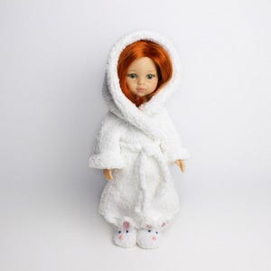 May include: A white hooded bathrobe with a plush texture, worn by a doll with red hair. The doll is wearing white bunny slippers.