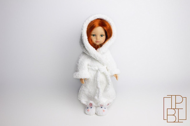 May include: A white hooded bathrobe with a plush texture, worn by a doll with red hair. The doll is wearing white bunny slippers.