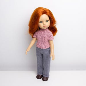 Pode incluir: Uma boneca com cabelo comprido e encaracolado vermelho vestindo um su&eacute;ter rosa de tric&ocirc; e cal&ccedil;as cinzas de tric&ocirc;. A boneca est&aacute; em p&eacute; sobre uma superf&iacute;cie branca.