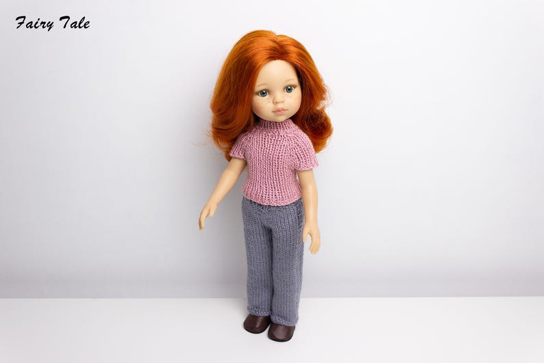 Pode incluir: Uma boneca com cabelo comprido e encaracolado vermelho vestindo um su&eacute;ter rosa de tric&ocirc; e cal&ccedil;as cinzas de tric&ocirc;. A boneca est&aacute; em p&eacute; sobre uma superf&iacute;cie branca.