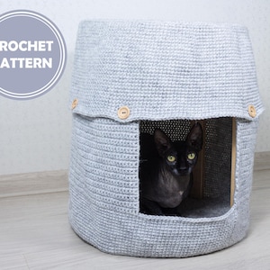 Puede incluir: Una cama para gatos de crochet gris con un gato negro dentro. La cama tiene una parte superior redondeada y una pequeña abertura para que el gato pueda entrar. La cama está hecha con un punto de crochet simple.