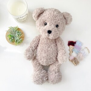 Pu&ograve; includere: Un orsacchiotto di peluche beige con occhi neri e naso nero. L'orso &egrave; seduto su una superficie bianca.