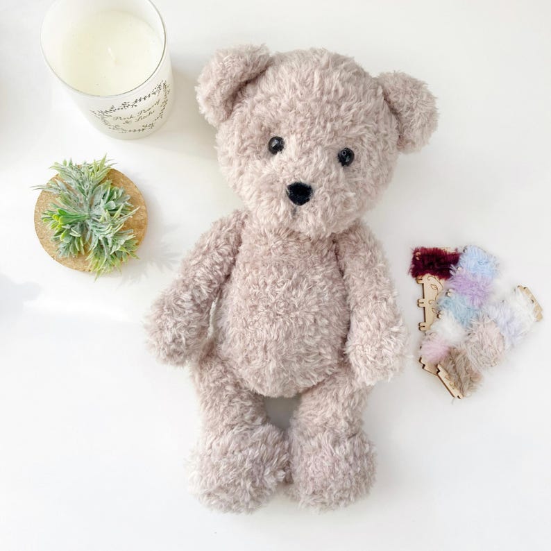 Pu&ograve; includere: Un orsacchiotto di peluche beige con occhi neri e naso nero. L'orso &egrave; seduto su una superficie bianca.