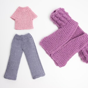 Pode incluir: Um conjunto de roupas de malha para uma boneca, incluindo um su&eacute;ter rosa, uma camisa rosa e cal&ccedil;as cinzas.