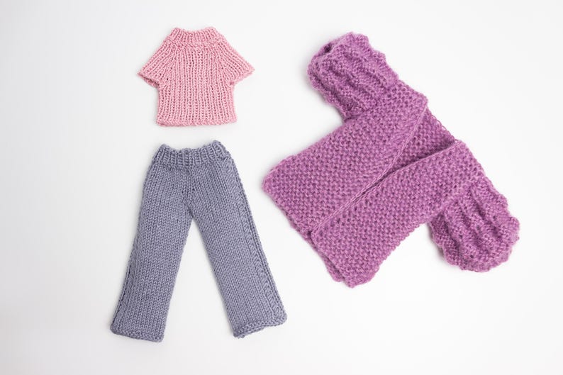 Pode incluir: Um conjunto de roupas de malha para uma boneca, incluindo um su&eacute;ter rosa, uma camisa rosa e cal&ccedil;as cinzas.