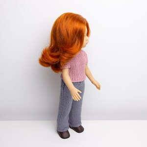 Pode incluir: Uma boneca com cabelo comprido, encaracolado e ruivo, vestindo uma blusa de malha rosa e cal&ccedil;as de malha cinzas. A boneca est&aacute; em p&eacute; sobre uma superf&iacute;cie branca.