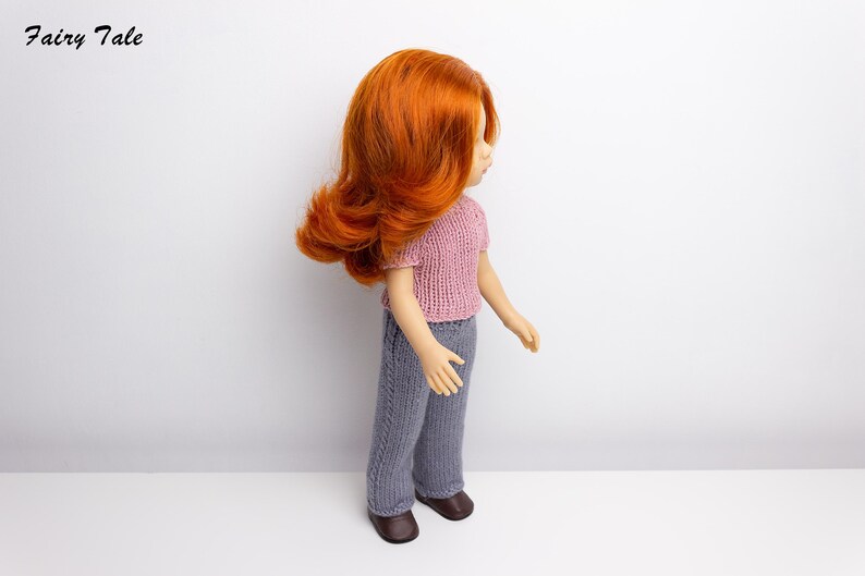 Pode incluir: Uma boneca com cabelo comprido, encaracolado e ruivo, vestindo uma blusa de malha rosa e cal&ccedil;as de malha cinzas. A boneca est&aacute; em p&eacute; sobre uma superf&iacute;cie branca.