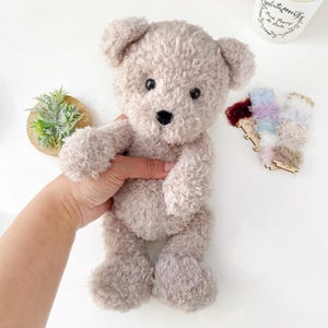 Pu&ograve; includere: Un orsacchiotto di peluche marrone chiaro con occhi neri e un naso nero. L'orso &egrave; in piedi su una superficie bianca.