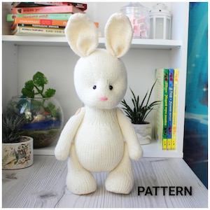 Könnte beinhalten: Ein weißes, gestricktes Stofftier in Form eines Hasen mit rosa Nase und schwarzen Augen. Der Hase steht auf einer Holzoberfläche mit einem Bücherregal im Hintergrund.