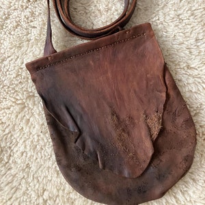 May include: A brown leather satchel with a long strap. The bag has a flap with a decorative edge and is worn and distressed.