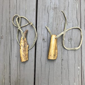 May include: Two pendants made from light brown, textured antler fragments, each attached to a thin, matching tan leather cord. The left pendant has a curved, tapered shape, while the right pendant is more cylindrical with a flat, angled top edge.