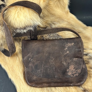 May include: A dark brown leather crossbody bag with a rectangular flap closure and a matching adjustable shoulder strap. The leather surface features a distressed, weathered texture with visible grain patterns and subtle lighter brown color variations.