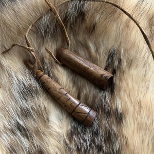 May include: Two brown wooden pendants suspended on thin leather cords. One pendant is cylindrical with a smooth finish, while the other is tapered and features etched horizontal and diagonal line patterns along its length.
