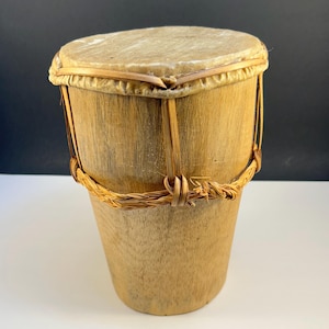 May include: A small, handcrafted bongo drum with a light brown wooden body and a natural hide drumhead. The drum is secured with woven rope and thin wooden supports, showcasing a rustic aesthetic. The drum is designed for musical performance.