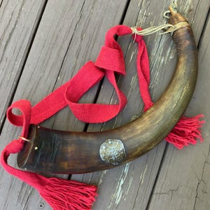 May include: A curved, brown horn with a red woven strap and tassels. The horn has a brass-colored metal accent and is attached to the strap with a metal ring. The horn is likely a decorative or functional item.