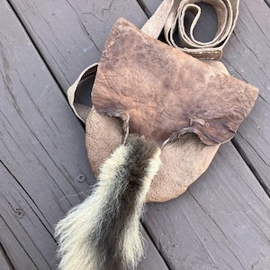 May include: A handmade leather shoulder bag with a flap closure. The bag is a light brown color with a darker brown flap. A long strap and a decorative tail with black and white fur are attached to the bag.