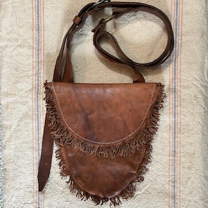 May include: A brown leather crossbody bag with a fringed bottom and a long adjustable strap. The bag has a flap closure and a buckle. The leather appears worn, giving it a vintage look. The bag is designed to be worn over the shoulder.
