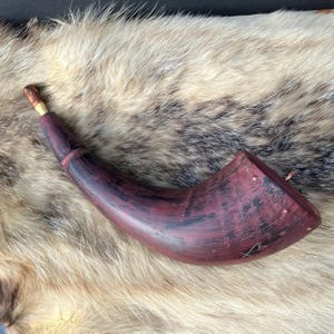 May include: A burgundy-colored powder horn with a curved shape and a wooden spout. The horn has dark accents and is decorated with small metal rivets. The background is a fur pelt.