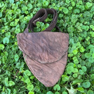 May include: A dark brown leather pouch with a triangular shape and a rounded flap closure. The bag features a textured, weathered surface and includes a long, matching leather strap with visible white stitching details at the connection point.