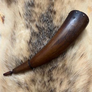 May include: A dark brown wooden horn with a turtle carving. The horn has a flared opening with metal studs and tapers to a narrow tip. The horn is likely used for ceremonial or decorative purposes.