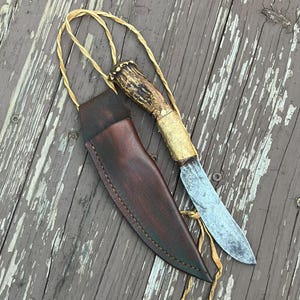 May include: A fixed-blade knife with a textured antler handle and a metallic bolster, accompanied by a dark brown leather sheath. A thin, light-colored cord is looped through the handle, providing a way to carry or hang the knife and sheath set.