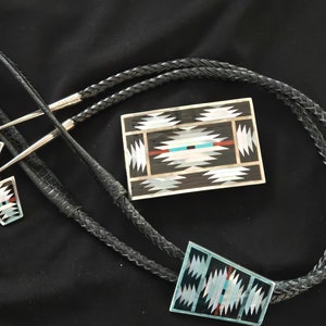 May include: A black leather bolo tie with a silver and turquoise bolo tie slide featuring a geometric pattern. The slide is made of inlaid mother of pearl and turquoise. The bolo tie is accompanied by a matching pair of earrings.