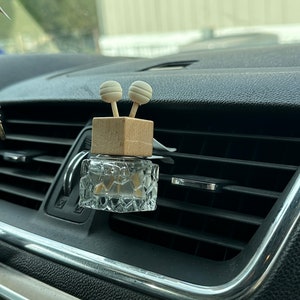 May include: A clear glass car air freshener with a wooden base and two white round diffusers. The air freshener is attached to a car's air vent.