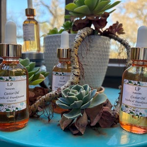 Puede incluir: Tres botellas de vidrio ámbar de aceite de ricino con cuentagotas dorados se exhiben. Cada botella tiene una etiqueta que dice "LP Naturals Castor Oil Jojoba & Frankincense". Las botellas están sobre una superficie turquesa, rodeadas de suculentas y una planta decorativa.