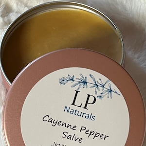 May include: A round, open, rose-gold tin of LP Naturals Cayenne Pepper Salve. The salve is a golden-yellow color. The label features the text "LP Naturals" and "Cayenne Pepper Salve" with a floral design. Net weight is 2 oz.