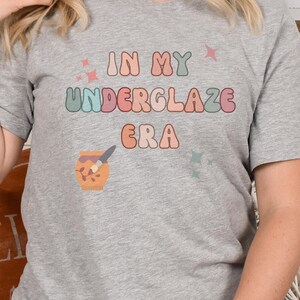 Può includere: Una maglietta grigia con il testo "In My Underglaze Era" in lettere colorate. Sotto il testo c'è una piccola illustrazione di un pennello e un barattolo di smalto.