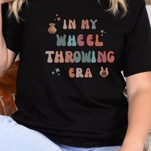 Può includere: T-shirt nero con la scritta "IN MY WHEEL THROWING ERA" in stile retrò colorato. Il design include un tornio da vasaio, stelle e un ferro di cavallo. La maglietta è abbinata a jeans azzurri e scarpe marroni.