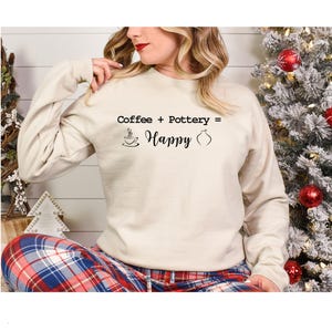 May include: A cream-coloured sweatshirt featuring the text "Coffee + Pottery = Happy" in black, with a coffee cup and vase graphic. The person is wearing plaid pyjama bottoms.