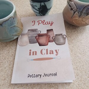 May include: A pottery journal titled "I Play in Clay" with a watercolor illustration of pottery vessels. The journal is open, with the text "Pottery Journal" at the bottom. Three ceramic mugs are in the background.