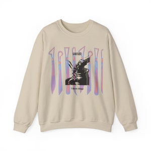 May include: A beige sweatshirt with a graphic print of a hand holding a knife and the text "CAUTION: I throw things". The graphic is surrounded by a row of knives with pink and blue gradients.