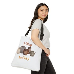 Può includere: Borsa tote bianca con tracolla nera. La borsa presenta la scritta "I Play in Clay" in marrone e un'illustrazione ad acquerello di ceramiche grigie e marroni. La borsa è portata a spalla.