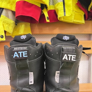 Può includere: Un paio di stivali da lavoro in pelle nera con un'etichetta blu e bianca che dice "ATE" su ogni stivale. Gli stivali hanno un'etichetta nera e bianca che dice "GORE-TEX" su ogni stivale. Gli stivali sono su una superficie di legno.