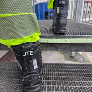 Könnte beinhalten: Schwarze Arbeitsschuhe mit einem gelben reflektierenden Streifen am Bein des Trägers. Die Schuhe tragen den Text "JTE" und "GORE-TEX® CROSS-TECH™".