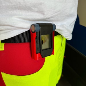 Puede incluir: Un dispositivo rojo y negro con una pequeña pantalla está sujeto a un cinturón negro. El dispositivo se lleva en la cintura de una persona.