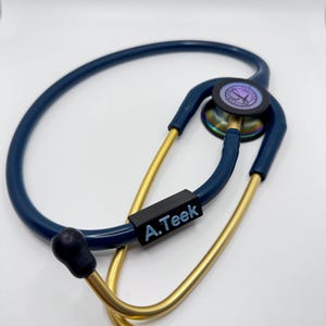 Könnte beinhalten: Ein blaues und goldenes Stethoskop mit einem schwarzen Namensschild, auf dem "A.Teek" steht.