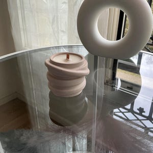 Peut inclure: Une bougie en béton rose avec une mèche en bois posée sur une table en verre. La bougie a un design texturé et ondulé.