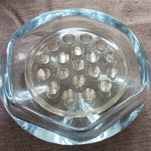 May include: A clear glass flower vase with a round base and a hexagonal top. The vase has a removable inner insert with multiple holes for holding flower stems.
