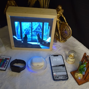 May include: A wooden frame with a light blue illuminated image of a house with two silhouettes of animals. The frame is on a white surface with a remote control, a smartwatch, a wireless charging pad, a smartphone with the time 19:20, a snow globe, and a wooden stand with a small plant.