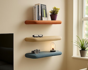 Puede incluir: Tres estantes flotantes en un salón moderno. El estante superior es naranja, con libros y una planta en maceta. El estante del medio es beige, con gafas. El estante inferior es azul, con un reloj y una pequeña lámpara.