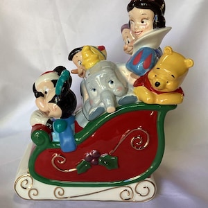 May include: A ceramic Christmas sleigh figurine featuring Mickey Mouse, Snow White, Dumbo, and Winnie the Pooh. The sleigh is red with green trim and gold accents. The characters are all smiling and appear to be enjoying a festive ride.