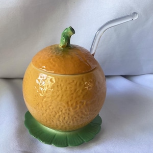 Könnte beinhalten: Ein keramik-orangenförmiges Glas mit Deckel und einem durchsichtigen Glaslöffel. Das Glas hat eine strukturierte orange Farbe mit einem grünen Stiel oben. Es steht auf einem grünen, blattförmigen Sockel. Der Löffel ist gebogen.