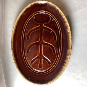 Könnte beinhalten: Ovale braune Keramikplatte mit glänzendem Finish. Die Platte hat ein erhabenes, stilisiertes Design in der Mitte. Der Rand hat einen helleren braunen, strukturierten Rand. Eine Vintage-Servierplatte.